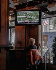 A person with short, light-colored hair is sitting in a cozy, wood-paneled interior of what appears to be a bar or cafe, watching a football game on a mounted TV screen. A warm-colored jacket is draped over their chair. The TV displays a live football match with a clear scoreboard and players on the field. The space is decorated with string lights, and an American flag is visible outside through a window next to some greenery.