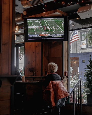 A person with short, light-colored hair is sitting in a cozy, wood-paneled interior of what appears to be a bar or cafe, watching a football game on a mounted TV screen. A warm-colored jacket is draped over their chair. The TV displays a live football match with a clear scoreboard and players on the field. The space is decorated with string lights, and an American flag is visible outside through a window next to some greenery.