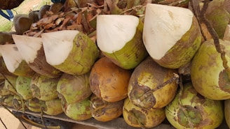 A collection of coconuts with their tops chopped off, exposing the white inner flesh. They are arranged in a large pile, showcasing their green and brown husks. Some dry leaves are visible in the background.