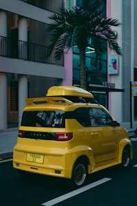 A compact, bright yellow car parked on a street against an urban backdrop with modern buildings. There is a palm tree nearby and visible reflections on the car's windows. The car has a boxy design with a roof box and custom plate, set in an environment that seems both urban and tropical.
