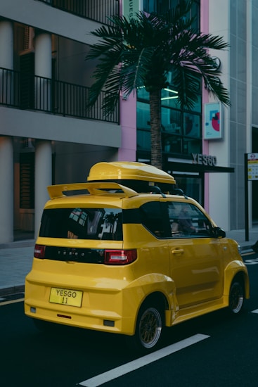 A compact, bright yellow car parked on a street against an urban backdrop with modern buildings. There is a palm tree nearby and visible reflections on the car's windows. The car has a boxy design with a roof box and custom plate, set in an environment that seems both urban and tropical.