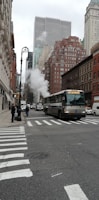 A bustling city street features a large 'Not In Service' bus traveling through an urban area. Tall buildings line the street on both sides, including distinctive skyscrapers in the background. Steam rises from a manhole, adding to the busy atmosphere. Pedestrians are visible on the sidewalks, and there are multiple vehicles on the road.