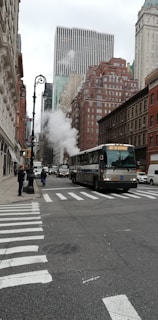 A bustling city street features a large 'Not In Service' bus traveling through an urban area. Tall buildings line the street on both sides, including distinctive skyscrapers in the background. Steam rises from a manhole, adding to the busy atmosphere. Pedestrians are visible on the sidewalks, and there are multiple vehicles on the road.