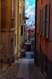A narrow cobblestone alley flanked by tall, colorful buildings with shuttered windows. Hanging planters with flowers decorate the space, adding a touch of greenery. Charming signs point towards a small hotel or shop further along the pathway. In the distance, a mountainous landscape is visible, under a bright, blue sky, providing a scenic backdrop to the quaint street.