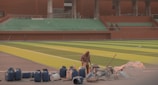 A construction worker in the foreground surrounded by blue barrels, construction materials, and tools. The background features a large empty stadium with green seating and a brightly colored green and yellow-striped field. The stadium structure includes modern architectural elements with a reddish-brown facade.