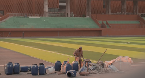 A construction worker in the foreground surrounded by blue barrels, construction materials, and tools. The background features a large empty stadium with green seating and a brightly colored green and yellow-striped field. The stadium structure includes modern architectural elements with a reddish-brown facade.