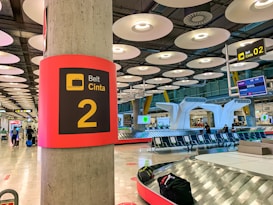 A modern airport baggage claim area with futuristic ceiling lights and a prominent sign displaying 'Belt Cinta 2'. The area has metallic baggage carousels, wide open spaces, and several people with luggage. Seating areas are visible, along with digital screens displaying flight information.