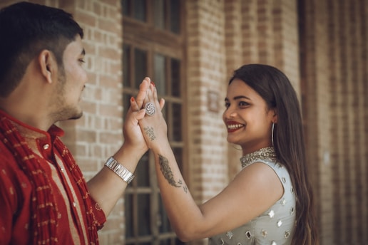 A smiling woman with long dark hair and hoop earrings is playfully high-fiving a man. She wears a sleeveless outfit adorned with decorative elements and has a tattoo and ring on her hand. The man, partially visible, wears a red traditional attire and a wristwatch. The background features a brick wall and a window with a warm ambiance.