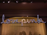 A sign displaying the words 'love pink' in a glowing, script-like font. The letters are adorned with small lights that give them a sparkling appearance. This sign is mounted on a softly lit wall, with ceiling lights above providing additional illumination.