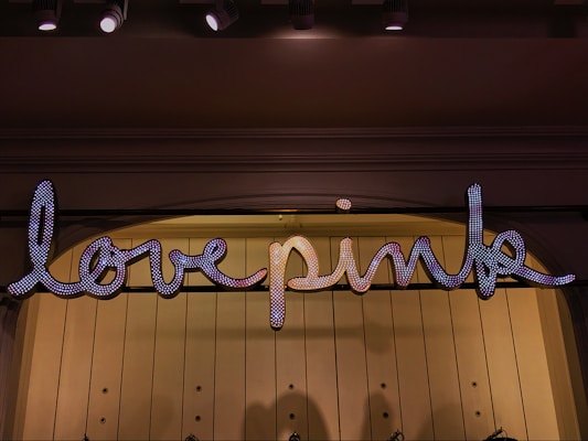 A sign displaying the words 'love pink' in a glowing, script-like font. The letters are adorned with small lights that give them a sparkling appearance. This sign is mounted on a softly lit wall, with ceiling lights above providing additional illumination.