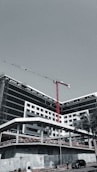 A large, modern building under construction with scaffolding and a tall red crane extending into the sky. The structure has a sleek design with black and white geometric patterns. A few people can be seen near the base, along with a parked car and a solitary palm tree.
