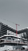 A large, modern building under construction with scaffolding and a tall red crane extending into the sky. The structure has a sleek design with black and white geometric patterns. A few people can be seen near the base, along with a parked car and a solitary palm tree.