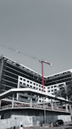 A large, modern building under construction with scaffolding and a tall red crane extending into the sky. The structure has a sleek design with black and white geometric patterns. A few people can be seen near the base, along with a parked car and a solitary palm tree.