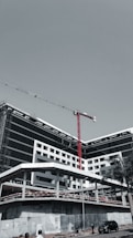 A large, modern building under construction with scaffolding and a tall red crane extending into the sky. The structure has a sleek design with black and white geometric patterns. A few people can be seen near the base, along with a parked car and a solitary palm tree.