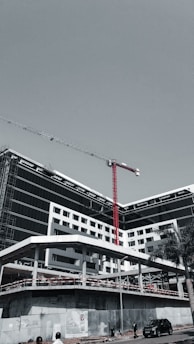 A large, modern building under construction with scaffolding and a tall red crane extending into the sky. The structure has a sleek design with black and white geometric patterns. A few people can be seen near the base, along with a parked car and a solitary palm tree.