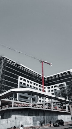 A large, modern building under construction with scaffolding and a tall red crane extending into the sky. The structure has a sleek design with black and white geometric patterns. A few people can be seen near the base, along with a parked car and a solitary palm tree.