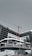 A large, modern building under construction with scaffolding and a tall red crane extending into the sky. The structure has a sleek design with black and white geometric patterns. A few people can be seen near the base, along with a parked car and a solitary palm tree.
