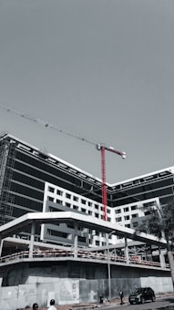 A large, modern building under construction with scaffolding and a tall red crane extending into the sky. The structure has a sleek design with black and white geometric patterns. A few people can be seen near the base, along with a parked car and a solitary palm tree.