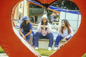 Three women are sitting together inside a heart-shaped frame. They appear to be engaged in a relaxed conversation, with one woman holding sunglasses and another wearing a hat. The surroundings include a grassy area and what looks like an outdoor setting with a few buildings in the background.