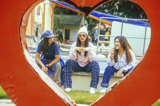 Three women are sitting together inside a heart-shaped frame. They appear to be engaged in a relaxed conversation, with one woman holding sunglasses and another wearing a hat. The surroundings include a grassy area and what looks like an outdoor setting with a few buildings in the background.
