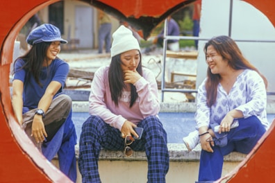 Three people are sitting together outdoors, framed by a large, red heart shape. They appear relaxed and are smiling, suggesting a friendly and cheerful atmosphere. The individual on the left wears a blue shirt and hat, the one in the middle is in a white beanie and pink sweatshirt, and the person on the right is in a white and blue patterned shirt.