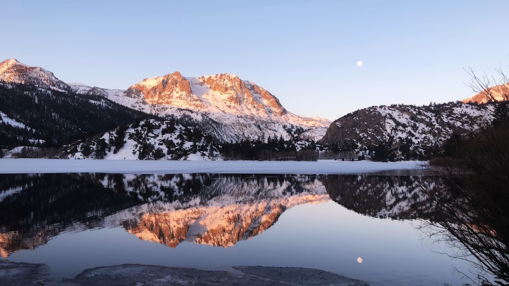 a lake surrounded by snow covered mountains with a moon in the sky