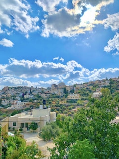 A scenic hillside community with a mix of modern and traditional houses, surrounded by lush greenery. The blue sky is dotted with fluffy white clouds, and sunlight peeks through, creating a serene atmosphere.