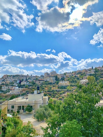 A scenic hillside community with a mix of modern and traditional houses, surrounded by lush greenery. The blue sky is dotted with fluffy white clouds, and sunlight peeks through, creating a serene atmosphere.