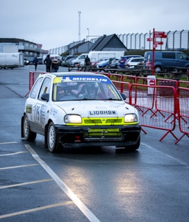 A rally car with a prominent white body and sponsor logos is navigating a wet asphalt track. The backdrop shows an overcast sky, several parked cars, people walking, and a fenced area with red barriers. The driver and co-driver inside the car are wearing helmets, and the vehicle has visible rally markings and numbers.