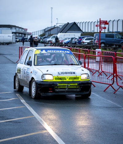 A rally car with a prominent white body and sponsor logos is navigating a wet asphalt track. The backdrop shows an overcast sky, several parked cars, people walking, and a fenced area with red barriers. The driver and co-driver inside the car are wearing helmets, and the vehicle has visible rally markings and numbers.