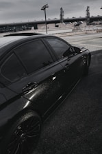 A sleek black car is parked on a rooftop or elevated parking area. The car's window is tinted, and the vehicle has shiny, well-maintained bodywork. In the background, industrial-looking buildings and lighting structures are visible under a gray, overcast sky.