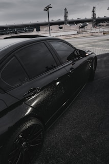 A sleek black car is parked on a rooftop or elevated parking area. The car's window is tinted, and the vehicle has shiny, well-maintained bodywork. In the background, industrial-looking buildings and lighting structures are visible under a gray, overcast sky.