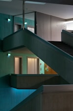 A multi-level interior space features intersecting staircases and illuminated exit signs, creating a geometric architectural design. The ambient lighting is dim, with turquoise hues casting a serene atmosphere across concrete textures.