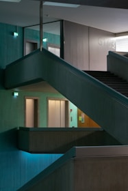 A multi-level interior space features intersecting staircases and illuminated exit signs, creating a geometric architectural design. The ambient lighting is dim, with turquoise hues casting a serene atmosphere across concrete textures.