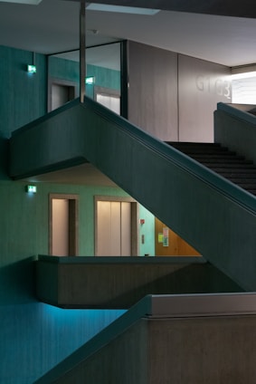 A multi-level interior space features intersecting staircases and illuminated exit signs, creating a geometric architectural design. The ambient lighting is dim, with turquoise hues casting a serene atmosphere across concrete textures.
