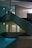 A multi-level interior space features intersecting staircases and illuminated exit signs, creating a geometric architectural design. The ambient lighting is dim, with turquoise hues casting a serene atmosphere across concrete textures.