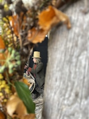 A person wearing a straw hat and coat is focused on weaving or crafting with natural materials. They are surrounded by dried leaves and branches, which frame the view.