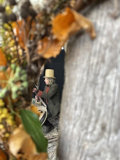 A person wearing a straw hat and coat is focused on weaving or crafting with natural materials. They are surrounded by dried leaves and branches, which frame the view.