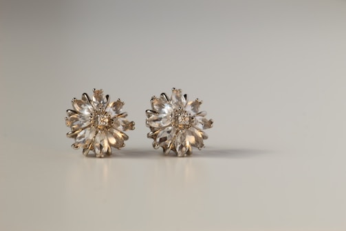Two elegant flower-shaped earrings are displayed on a plain background. The earrings are adorned with clear, sparkling gemstones set in a radial pattern, resembling a blossoming flower. Each earring has intricate settings that catch the light, creating a sense of luxury.