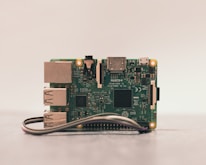 A close-up of a Raspberry Pi board placed on a surface. The green circuit board features various visible components such as USB ports, HDMI port, and several microchips. A ribbon cable is partially visible at the bottom.