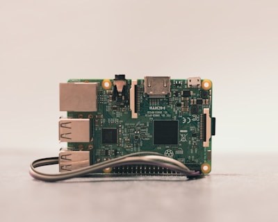 A close-up of a Raspberry Pi board placed on a surface. The green circuit board features various visible components such as USB ports, HDMI port, and several microchips. A ribbon cable is partially visible at the bottom.