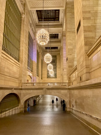 An elegant stone interior with high ceilings decorated with ornate chandeliers. The architecture features large, arched windows and spacious corridors. Several people are walking through the grand hallway, creating a sense of movement in the otherwise tranquil space.