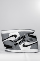 A pair of grey and white high-top sneakers with black accents, featuring the iconic Nike swoosh and Air Jordan logo. The sneakers are positioned sideways, stacked neatly on top of each other against a plain background.