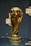 A golden trophy is displayed, featuring intricate sculpted details and a globe at the top. It is set on a dark background with reflective surfaces around it.