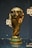 A golden trophy is displayed, featuring intricate sculpted details and a globe at the top. It is set on a dark background with reflective surfaces around it.