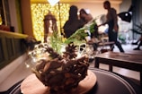 A wooden decorative centerpiece with pine cones and greenery is placed on a table. In the background, warm yellow fairy lights create a cozy, festive ambiance. There are indistinct silhouettes of people engaging in activities, suggesting a social gathering or event.