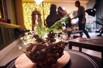 A wooden decorative centerpiece with pine cones and greenery is placed on a table. In the background, warm yellow fairy lights create a cozy, festive ambiance. There are indistinct silhouettes of people engaging in activities, suggesting a social gathering or event.