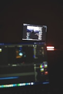 A dimly lit room with two computer screens displaying editing software. The foreground monitor shows various color and timeline adjustments, while the background monitor displays a video of a person in front of a large landscape. The ambiance is focused and technology-oriented.
