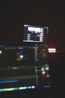 A dimly lit room with two computer screens displaying editing software. The foreground monitor shows various color and timeline adjustments, while the background monitor displays a video of a person in front of a large landscape. The ambiance is focused and technology-oriented.