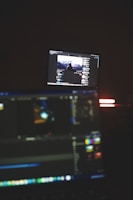 A dimly lit room with two computer screens displaying editing software. The foreground monitor shows various color and timeline adjustments, while the background monitor displays a video of a person in front of a large landscape. The ambiance is focused and technology-oriented.